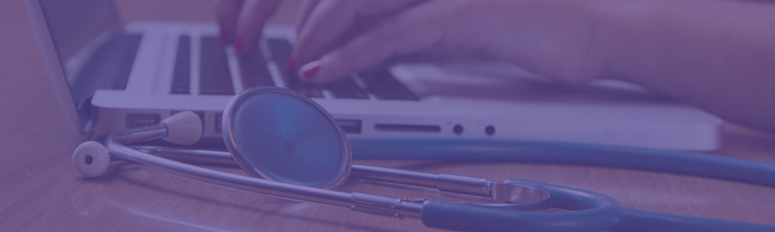 A stethoscope on a desk next to a laptop with hands typing