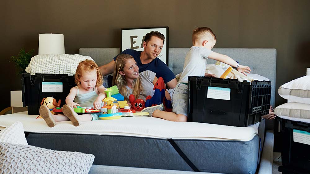 A young family of four on a unmade bed and unpacking moving home boxes