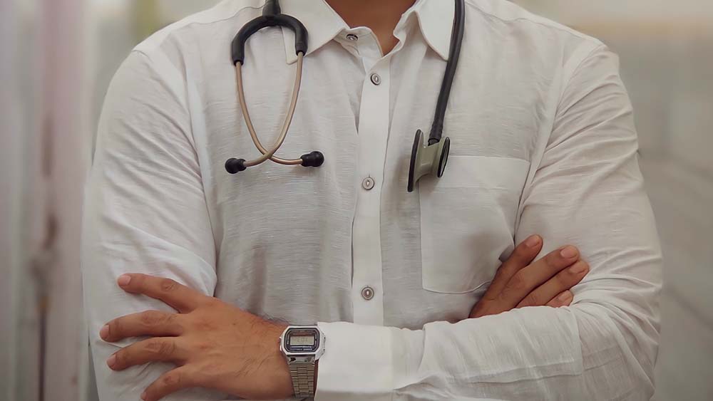 A stethoscope around the neck of a GP with crossed arms and white shirt