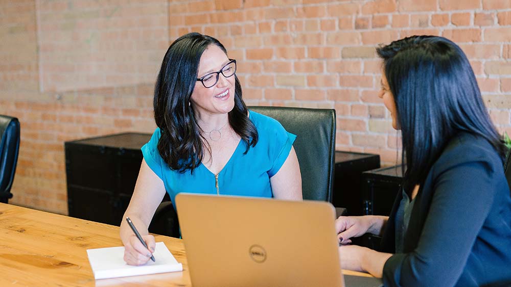 Two women with black hair talking at an office table with notepad and laptop