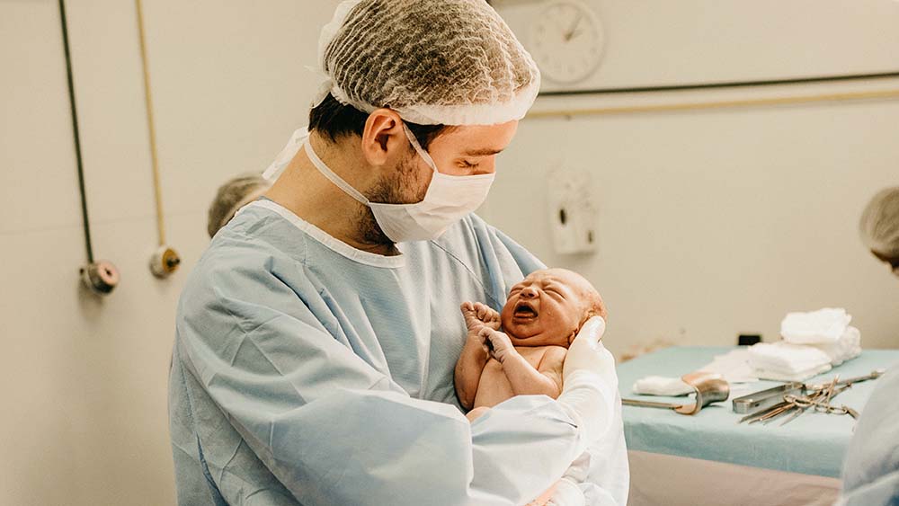 A new born baby being held by a man in hospital scrubs, hair net and face mask