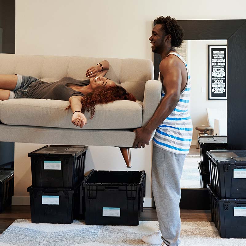 Man lifting a sofa with lady lying on the sofa and laughing.