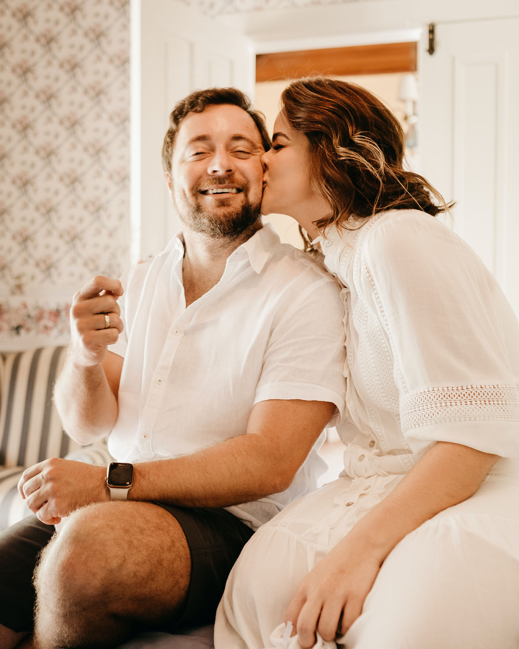 A bearded man being kissed on the cheek by his wife. Both are sitting and dressed in white shirts.