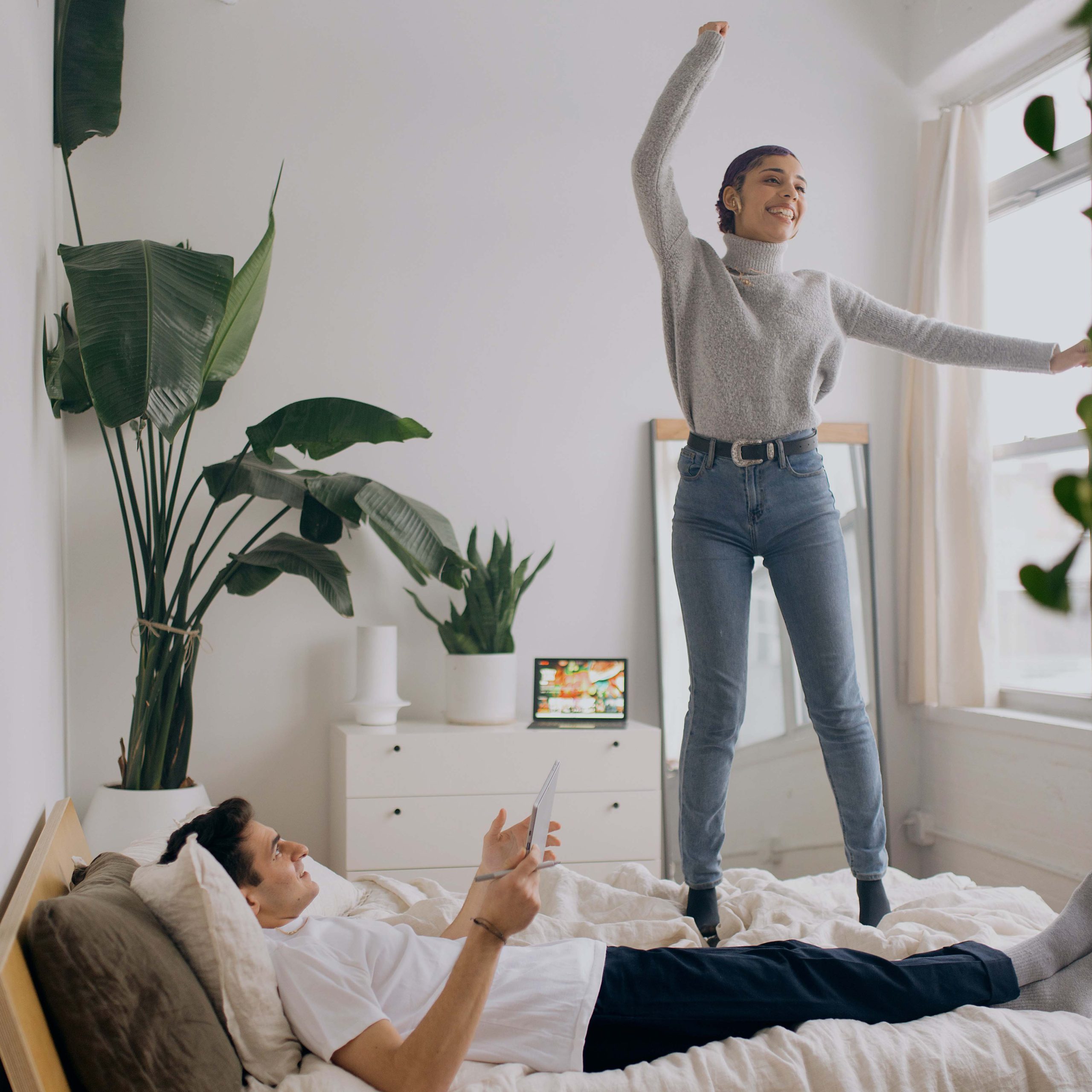 Young lady bouncing on her bed with partner watching and smiling