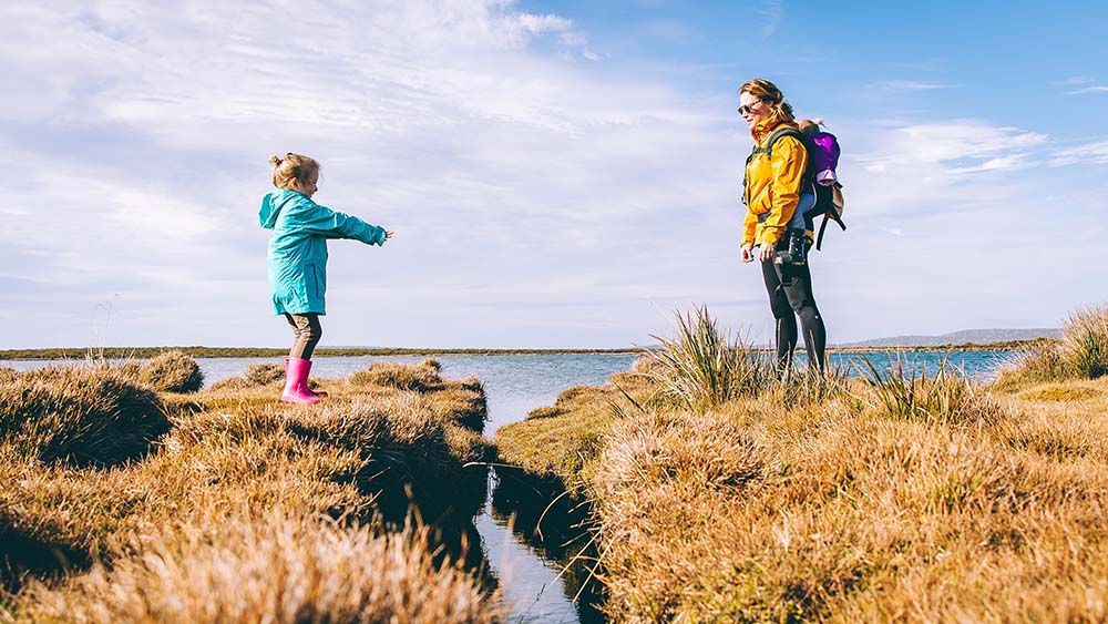 A child in a green coat holds out hand to a woman with a yellow rain coat and backpack to cross a gully full of water