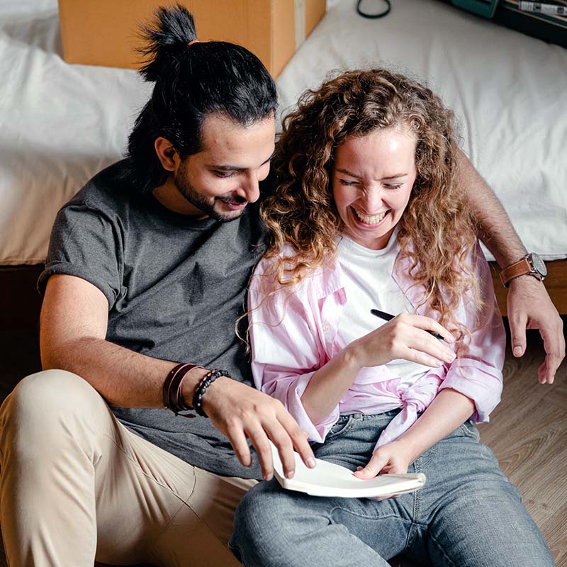A couple sitting against a bed laughing and looking at a note